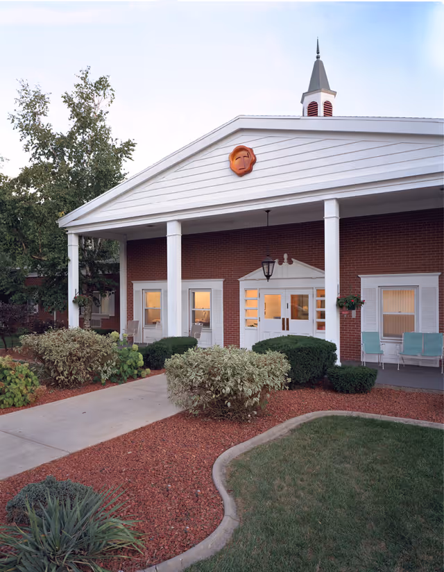 Exterior view of St. Mary Healthcare Center showing a brick building with white columns and a peaked roof with a small steeple. There are shrubs and landscaped areas with red mulch and a concrete walkway leading to the entrance. The entrance has double doors with windows and outdoor seating on the porch.