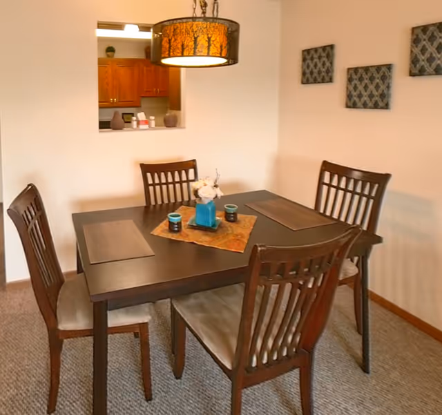 Dining area with a dark wood table and four chairs beneath a tree-patterned pendant light and a kitchen pass-through in the background.