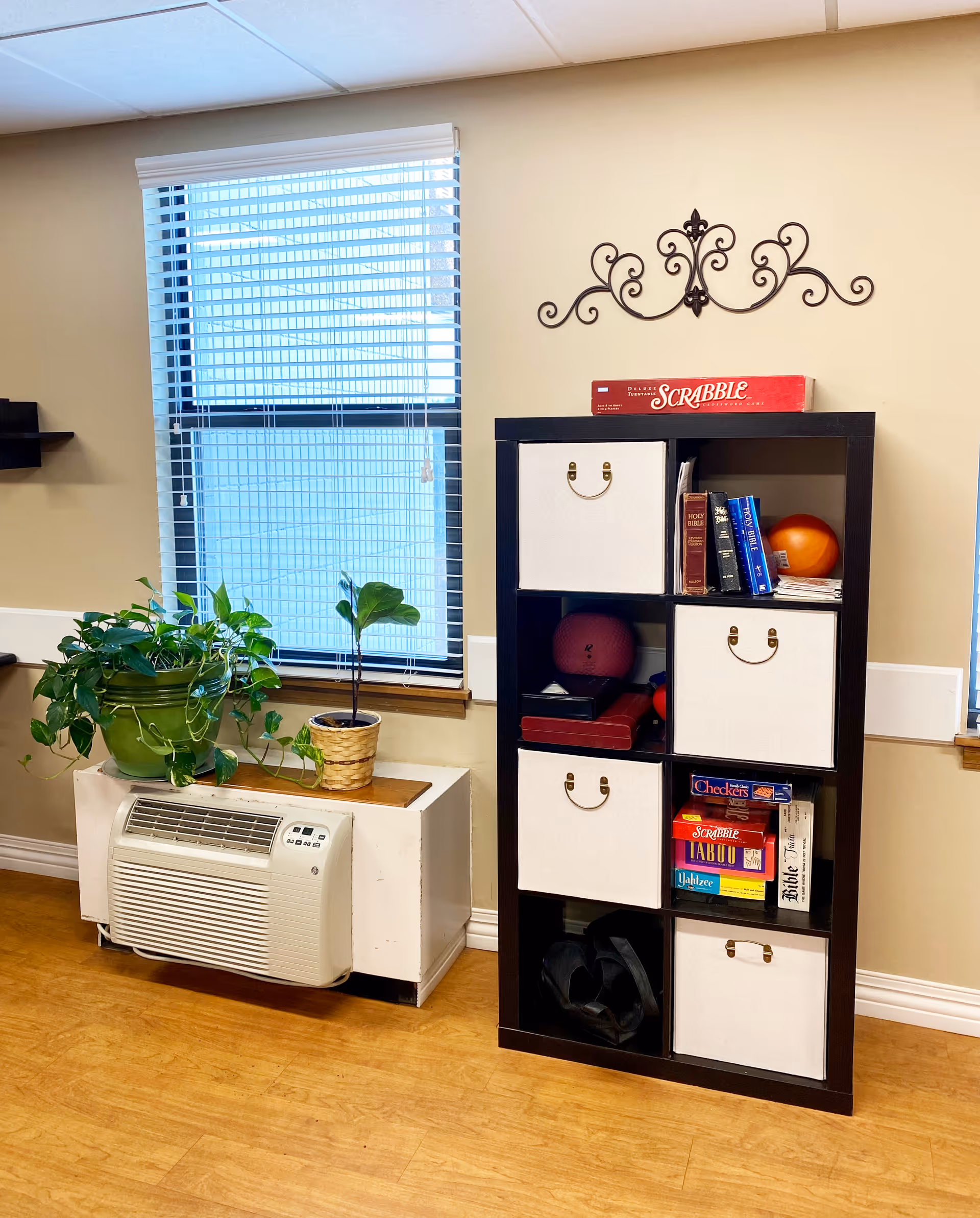 Interior corner of a room with a window covered by white blinds, a white air conditioning unit below the window, and two potted plants on top of a white radiator cover. Next to it is a black shelving unit with beige fabric storage bins, books, board games including Scrabble, Checkers, Taboo, and Yahtzee, and some exercise equipment. A decorative metal wall art piece is mounted above the shelving unit.