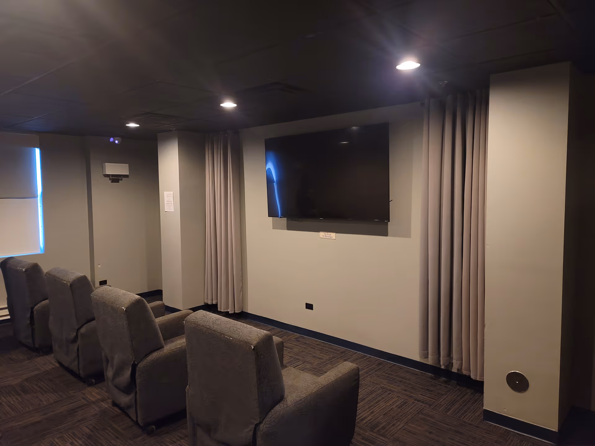 Small screening room with a wall-mounted TV, rows of upholstered recliner chairs, and closed curtains.