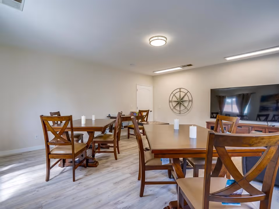 A well-lit room with wooden tables and chairs arranged neatly, each table having white candles on top. The room has light-colored walls and flooring, a large flat-screen TV mounted on a wooden cabinet, and a decorative wall piece resembling a compass rose. There is a door in the background and a window with curtains reflected in the TV screen.