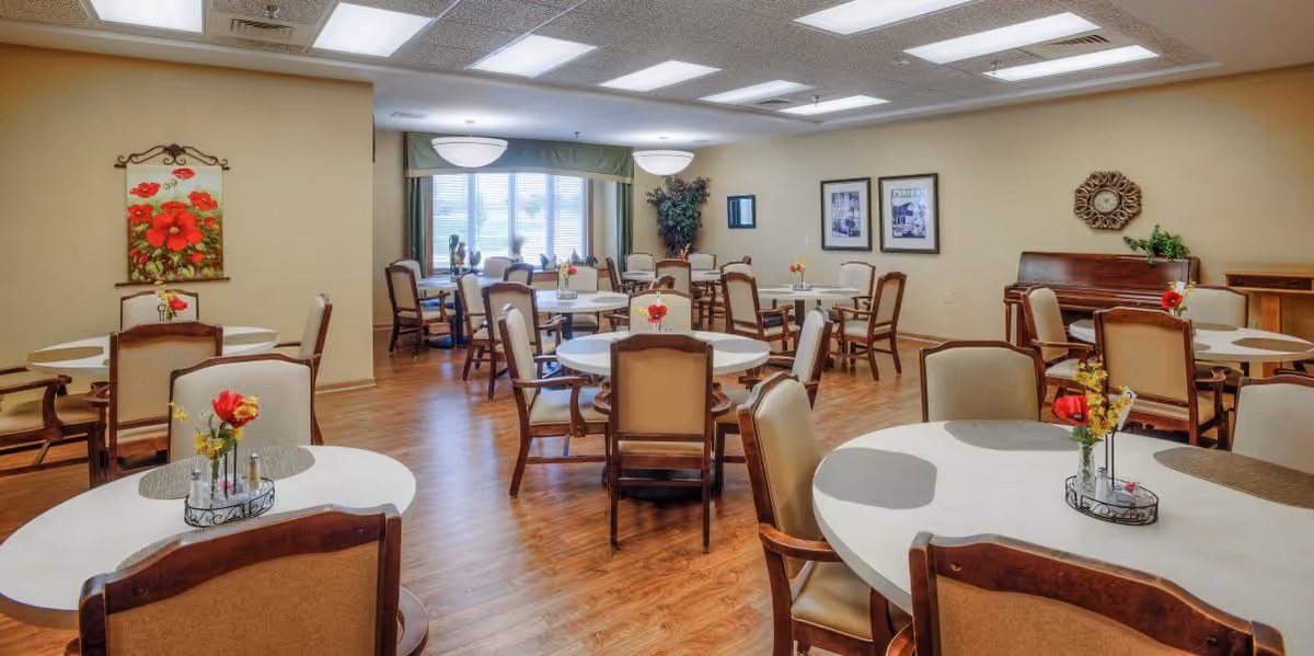 A bright dining room with multiple round tables and cushioned chairs arranged neatly. Each table has a small floral centerpiece. The room has wooden flooring, beige walls adorned with framed pictures and a decorative mirror, and large windows with green valances letting in natural light.