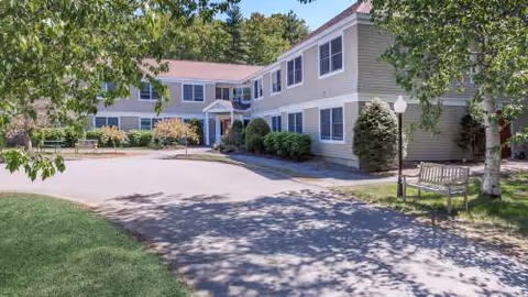 Exterior view of Merry Gardens senior living facility with trees, walkway, and benches.