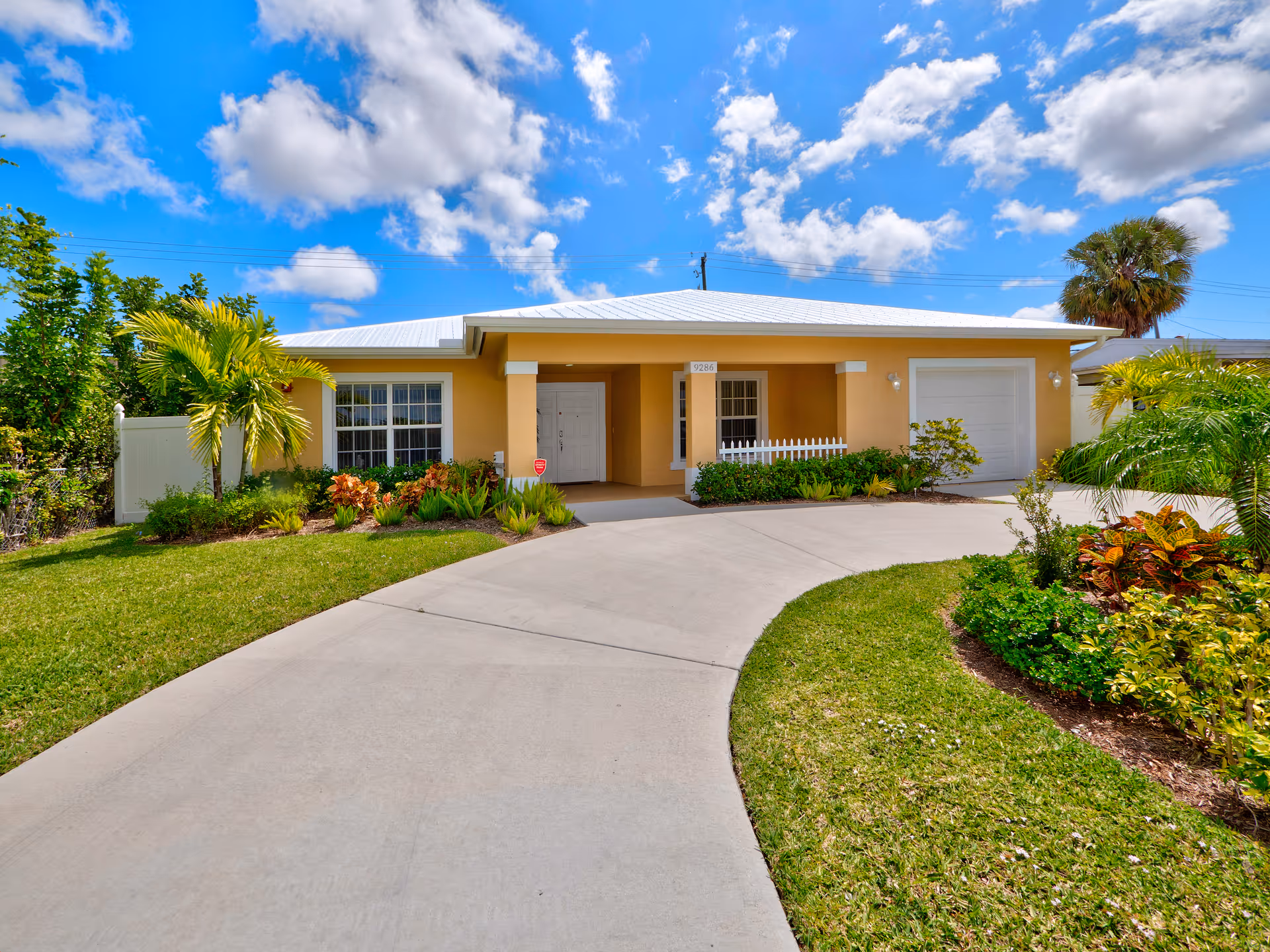 Front exterior view of a single-story yellow house with a white roof, a curved concrete driveway, green lawn, and landscaped garden beds with various plants and palm trees under a blue sky with scattered clouds.