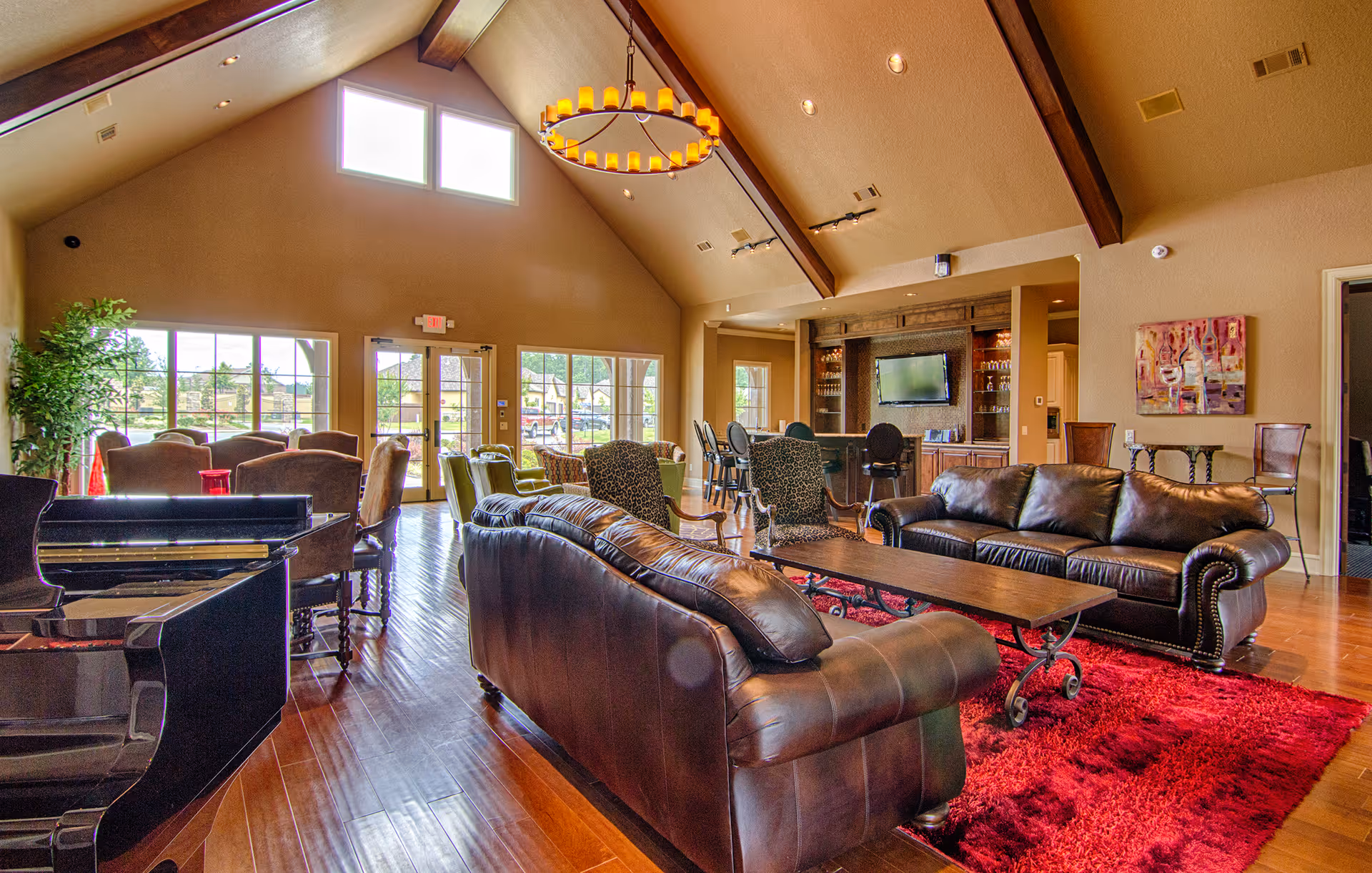 Spacious living room area with high vaulted ceilings and exposed wooden beams. The room features two dark leather sofas, a wooden coffee table on a red rug, several patterned armchairs, a grand piano, and a bar area with stools and a mounted TV. Large windows and glass doors allow natural light to fill the space.