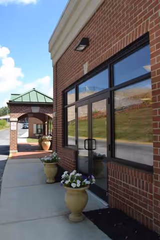 Brick senior living facility exterior with large windows, an entrance, and potted flowers.