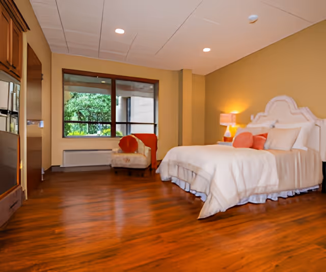 Spacious bedroom with a large bed, an armchair by a window, wooden floors and a bedside lamp.