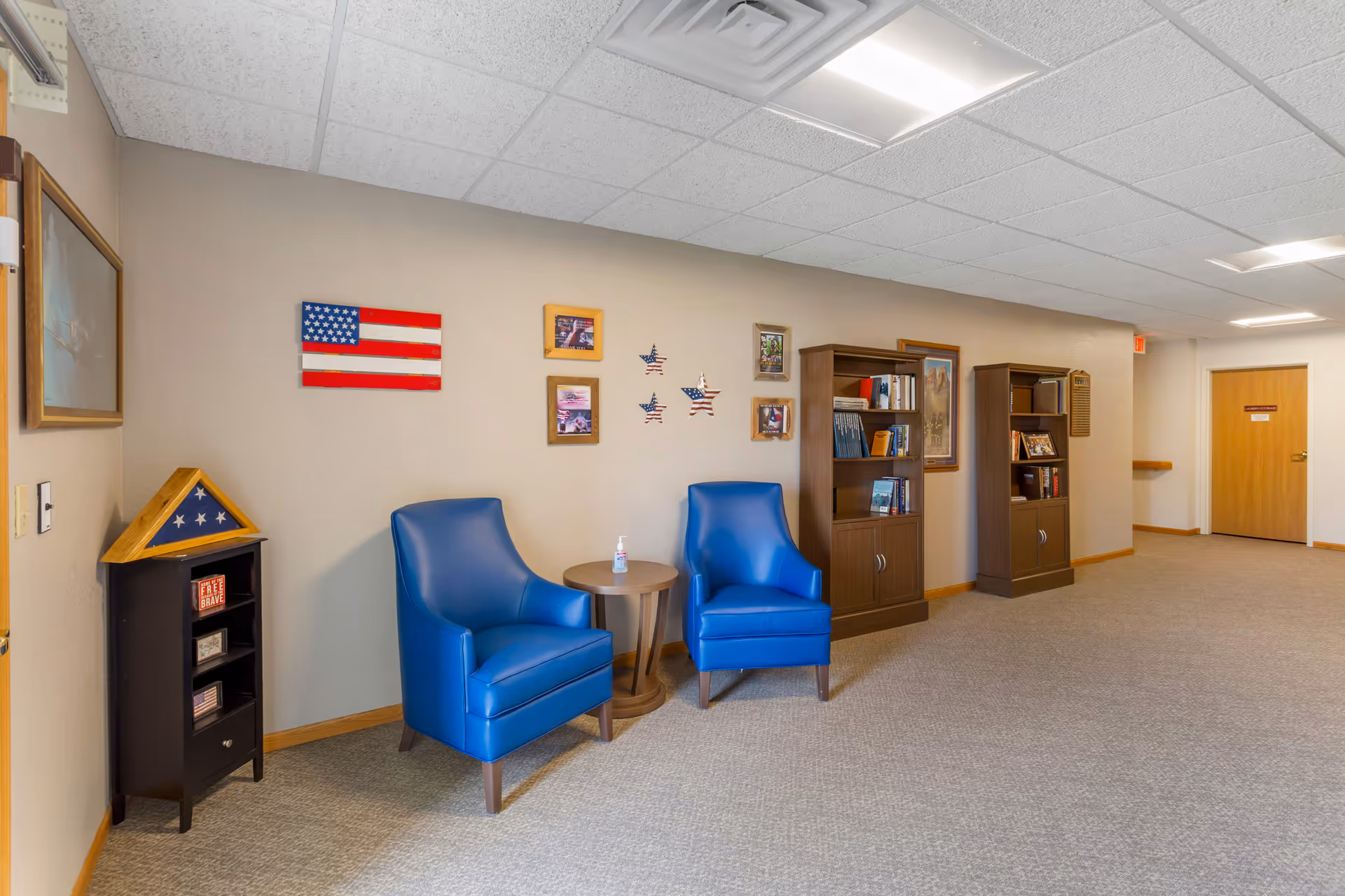 A hallway area in South Grove Lodge Senior Living with two blue armchairs and a small round wooden table between them. The wall behind features an American flag, framed pictures, and star-shaped decorations. There are two wooden bookshelves filled with books and framed photos. The floor is carpeted, and the ceiling has white tiles with fluorescent lighting.