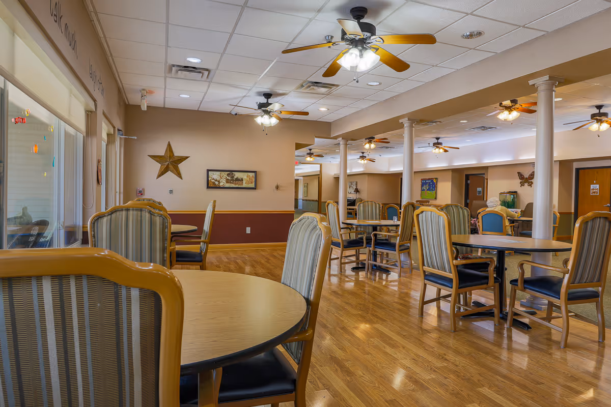 A spacious dining area in an assisted living facility with multiple round tables and cushioned chairs arranged neatly. The room features wooden flooring, beige walls with decorative elements like a star and framed artwork, ceiling fans with lights, and large windows on the left side letting in natural light.
