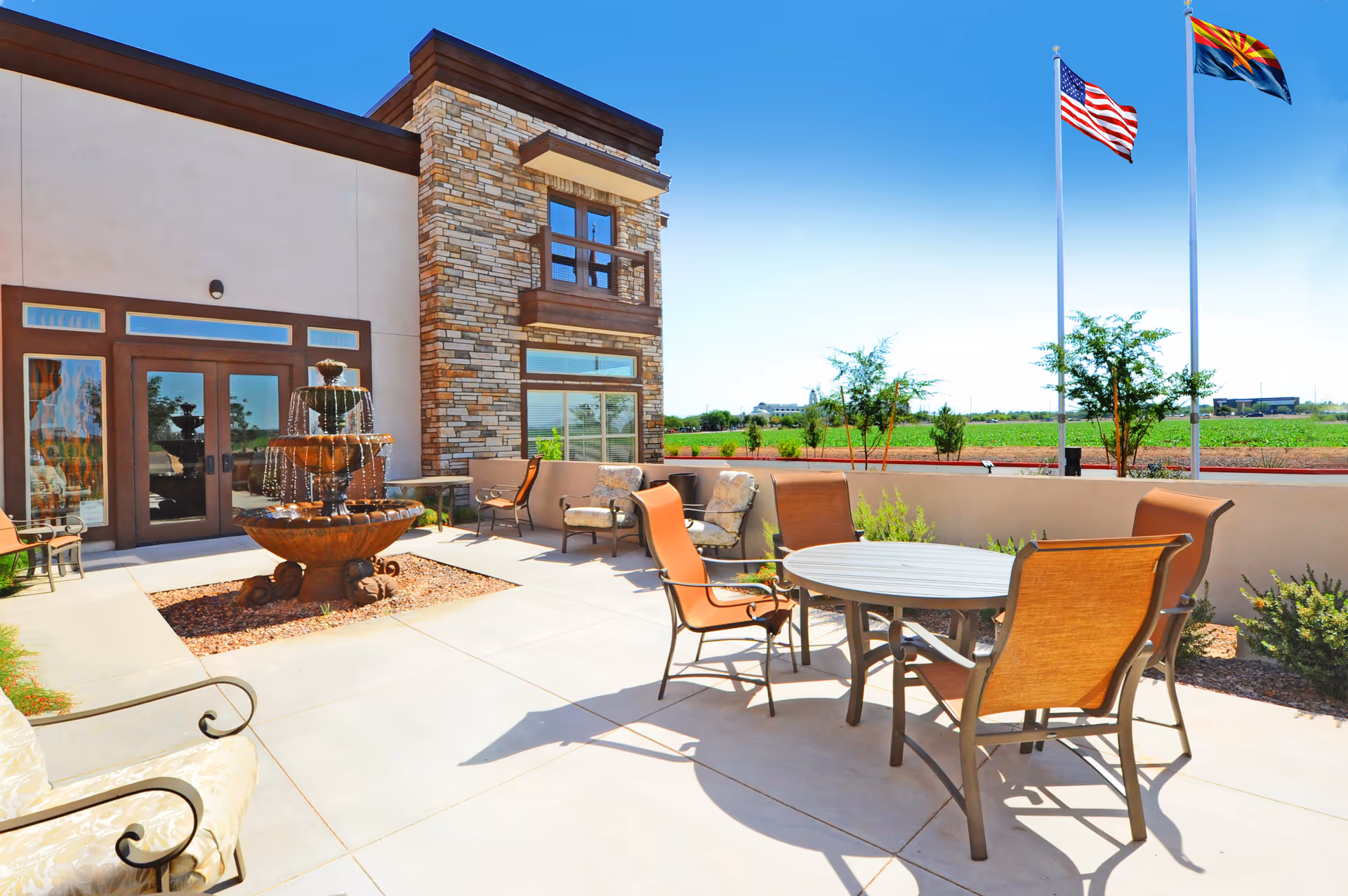 Outdoor patio area at Sky Ridge Senior Living with a round table and four chairs, cushioned armchairs, a three-tiered water fountain, and two flagpoles flying the American and Arizona state flags under a clear blue sky.
