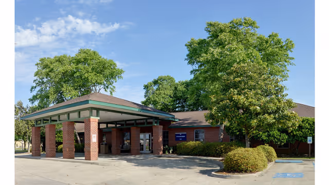 Single-story brick nursing home with a covered drive-up entrance, landscaped trees, and parking spaces.
