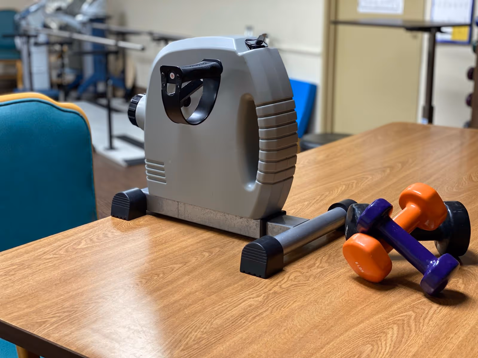A small exercise pedal machine and three colorful dumbbells (orange, purple, and black) placed on a wooden table in a room with exercise equipment and chairs in the background.