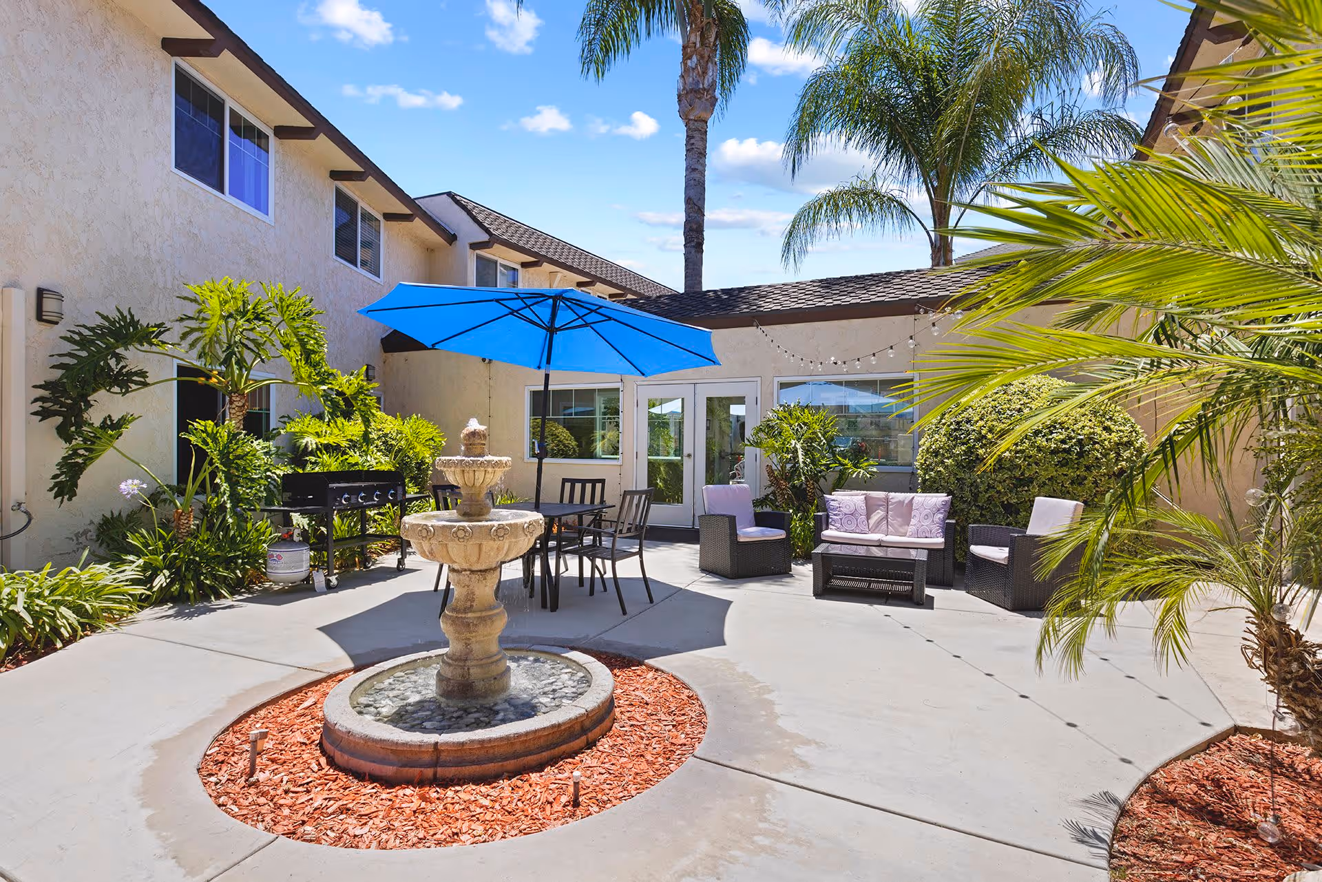 Courtyard patio with a central stone fountain, outdoor dining set under a blue umbrella, lounge seating, and palm trees.