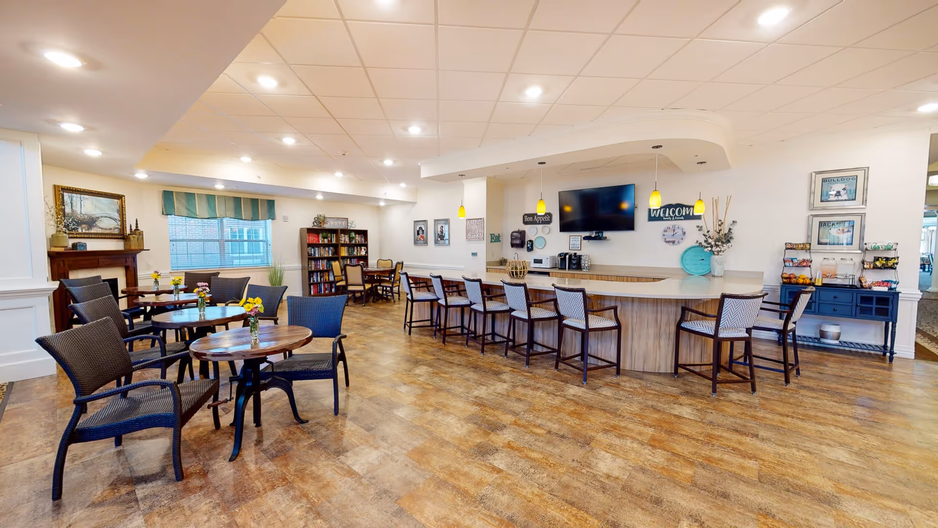 A spacious and well-lit common area with multiple round tables and chairs arranged for seating. There is a long counter with high chairs, pendant lights hanging above, and a wall-mounted TV. The room features wooden flooring, a bookshelf, framed pictures on the walls, and decorative items including flowers on the tables and a fireplace on the left side.