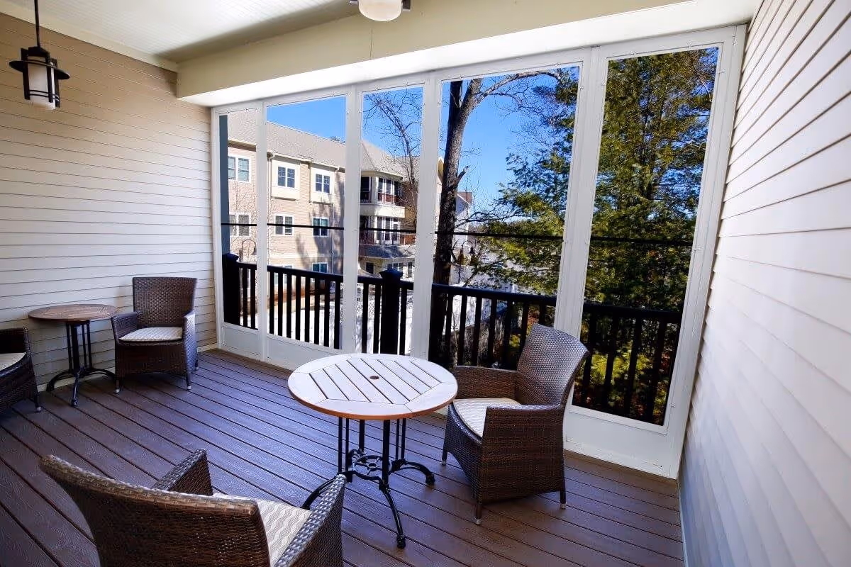 A screened-in porch with three wicker chairs and two round wooden tables. The porch has wooden flooring and white paneled walls. Outside the porch, there are trees and a view of a neighboring building under a clear blue sky.