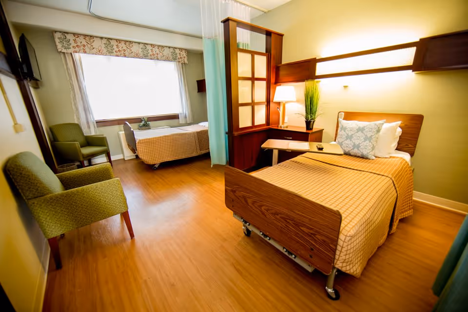 A well-lit room with two hospital-style beds separated by a wooden partition with frosted glass panels. Each bed has a checkered bedspread and pillows. There are two green upholstered chairs near a large window with floral valance and sheer curtains. A bedside table with a lamp and a potted plant is next to one of the beds. The floor is wooden and the walls are painted light green.