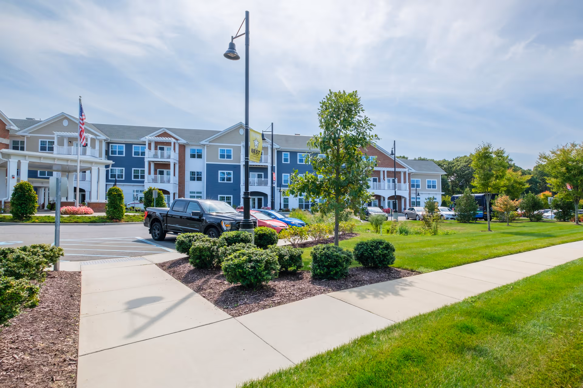 Exterior view of a senior living facility with a three-story building featuring balconies, an American flag, a parking lot with several cars, green lawns, trees, and a clear blue sky.