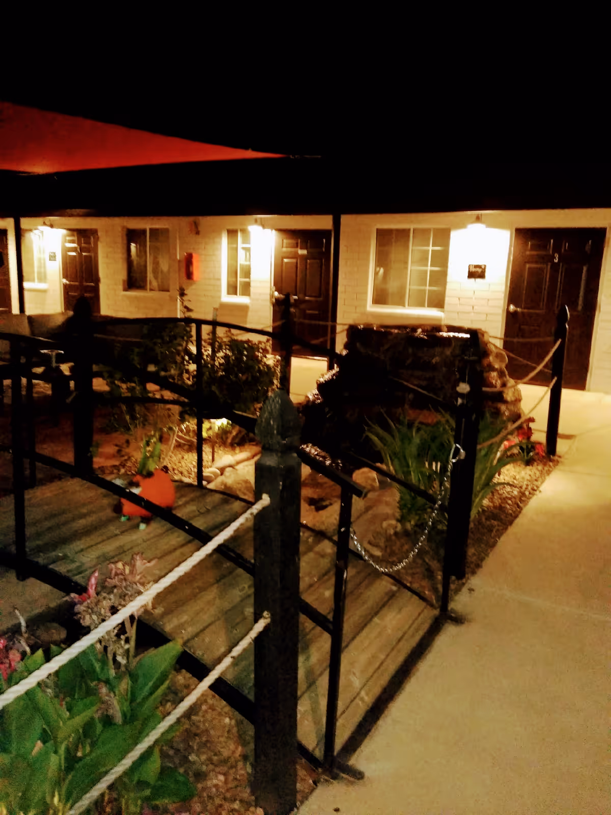 Nighttime view of an outdoor area at a senior living facility with a small wooden bridge over a landscaped garden. The area is illuminated by wall-mounted lights near several dark brown doors and windows on a light-colored building exterior. Plants and decorative rocks surround the bridge and walkway.