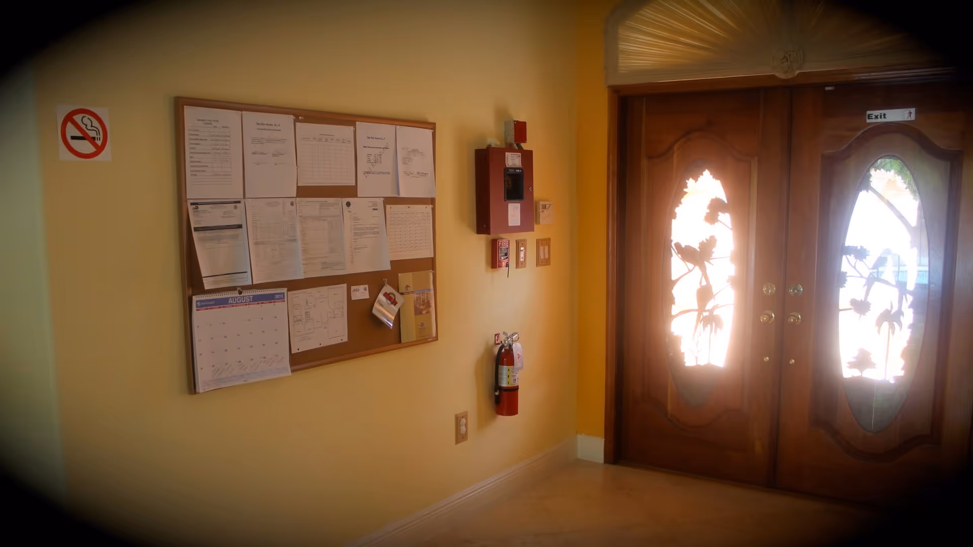 Interior view of a hallway corner with a bulletin board displaying various papers and a calendar on a yellow wall. There is a no smoking sign above the bulletin board, a fire extinguisher mounted on the wall, and double wooden doors with decorative glass panels labeled with an exit sign.