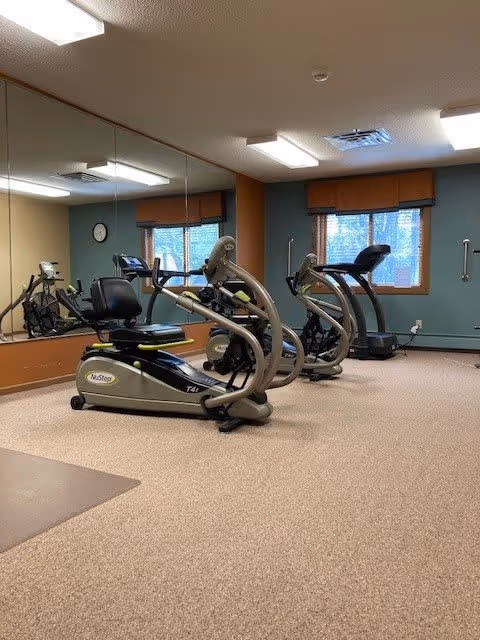 Interior view of a fitness room with exercise equipment including a recumbent bike and a treadmill. The room has carpeted flooring, large mirrors on one wall, windows with brown valances, and fluorescent ceiling lights.
