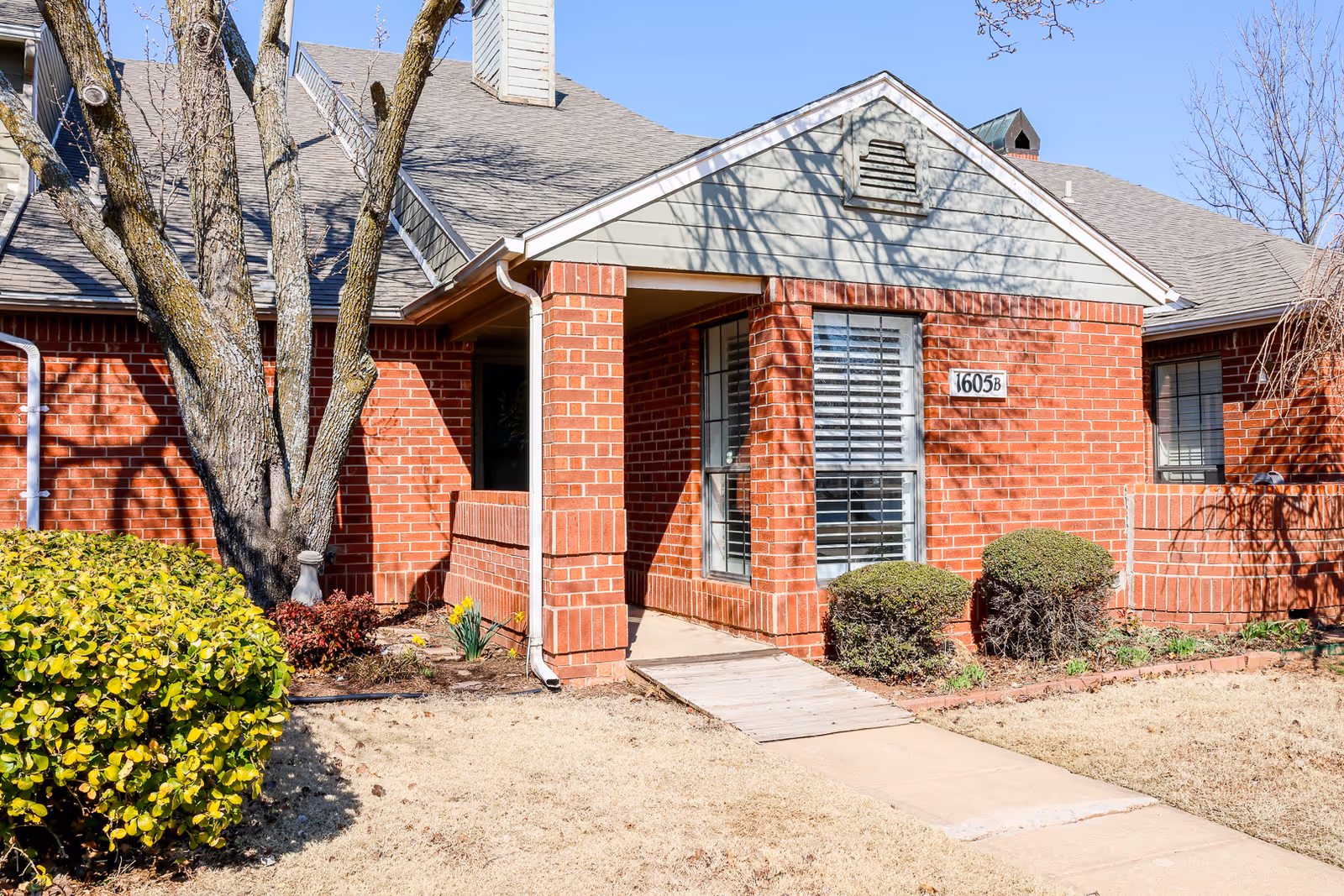 Front entrance of a red brick senior living unit marked 1605B with a small covered porch, shrubs, and a walkway.