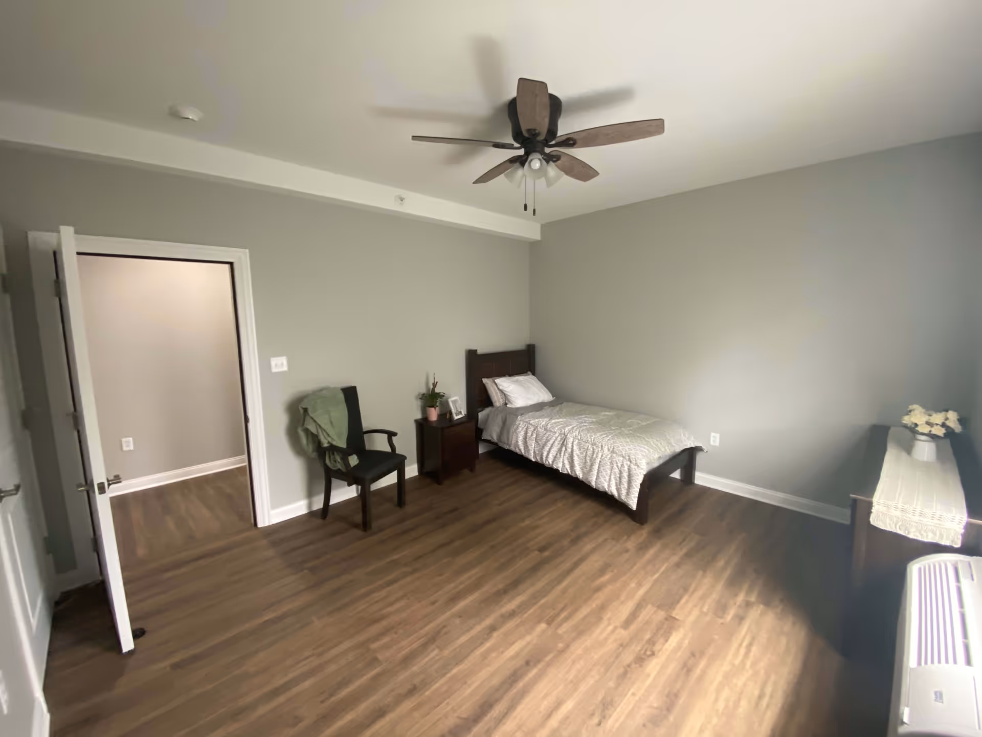 A simple bedroom with a single bed against the wall, a nightstand with a plant and picture frame, a wooden chair with a green cloth draped over it, a ceiling fan with lights, and a dresser with a vase of flowers. The room has wooden flooring and light gray walls.