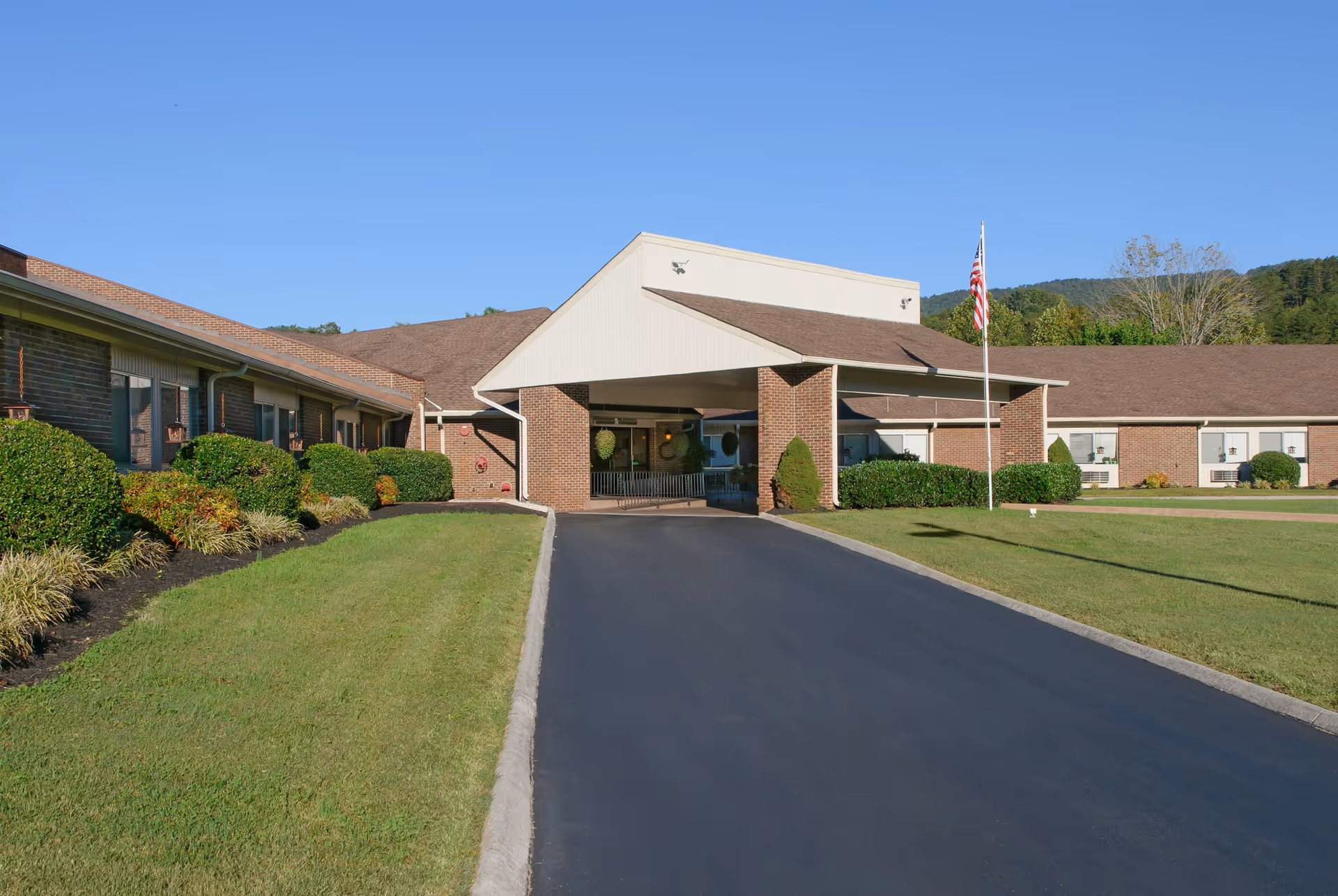 Exterior view of Ridgeview Terrace of Life Care building with a covered entrance, a paved driveway, well-maintained green lawns, bushes, and an American flag on a flagpole under a clear blue sky.