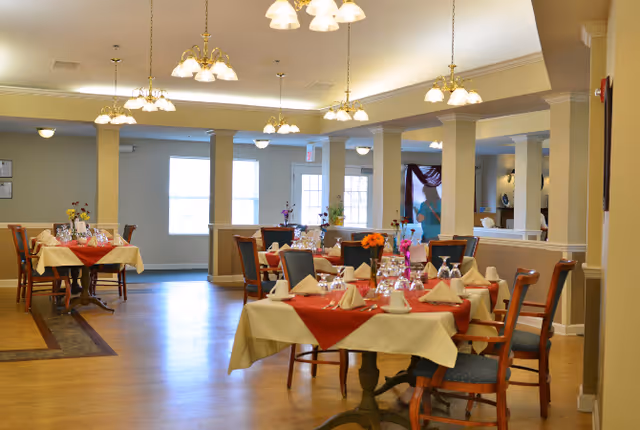 A spacious dining room with several tables covered in cream tablecloths and red table runners, each set with folded napkins, glasses, and small floral centerpieces. The room has wooden floors, multiple hanging light fixtures, and large windows allowing natural light to enter.
