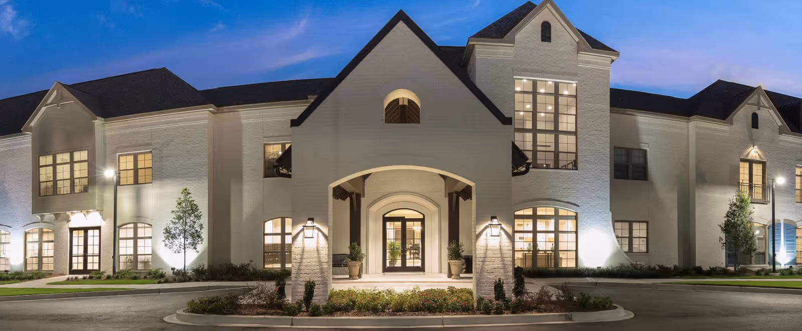 The Blake at Edgewater building exterior at dusk, showing a large, well-lit entrance with white brick walls, multiple windows, and a landscaped area in front.