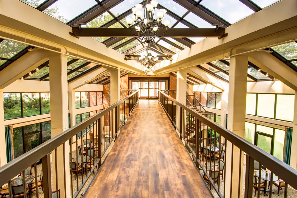 Interior view of a senior living facility with a wooden walkway on the second floor, metal railings, large windows, and a glass ceiling allowing natural light to fill the space. Below the walkway are seating areas with tables and chairs surrounded by large windows and greenery outside.