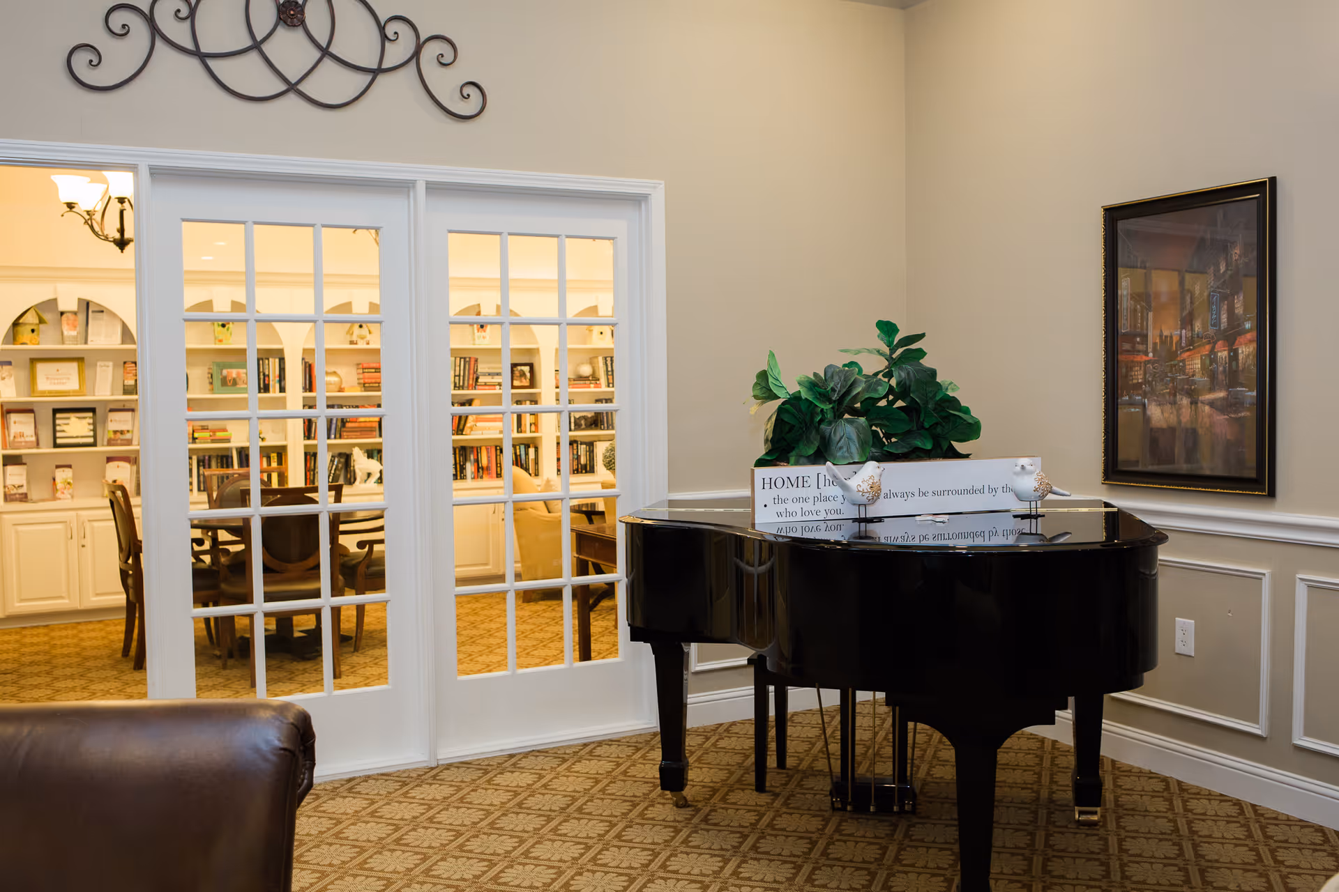 Interior common room with a black grand piano topped with a plant and sign, and glass-paned doors opening to a bookshelf-lined seating area.