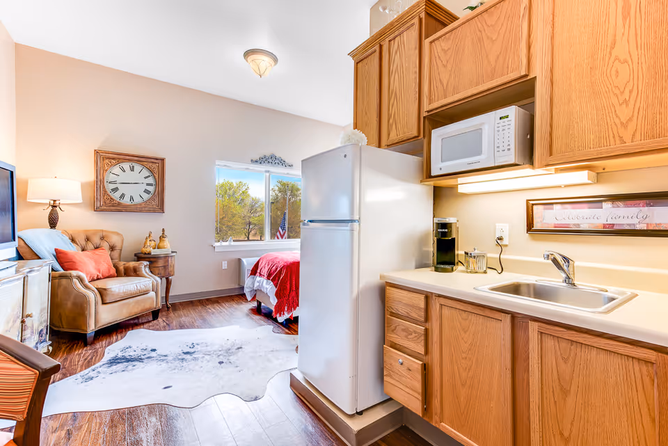 A cozy senior living studio apartment featuring a small kitchen area with wooden cabinets, a white refrigerator, microwave, and sink. The living space includes a comfortable armchair with a red pillow, a side table with decorative items, a wall clock, and a window with a view of trees and an American flag outside. A bed with a red blanket is partially visible near the window.