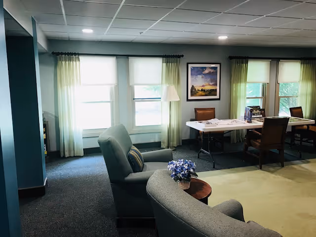 Bright communal living area with upholstered armchairs, a small round table with flowers, and folding tables and chairs by windows with green curtains.