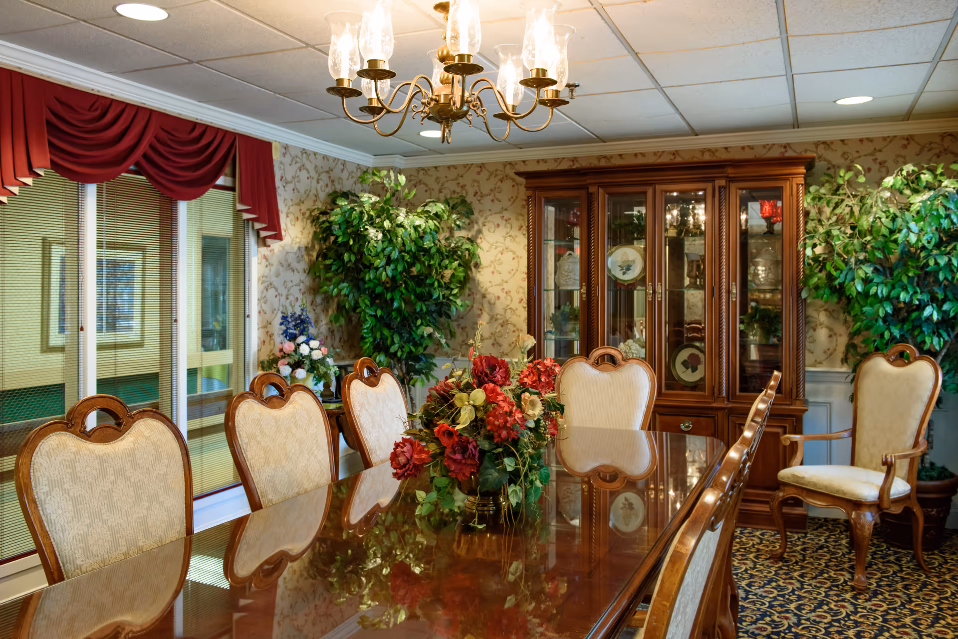 A formal dining room with a polished wooden table surrounded by upholstered chairs. A floral centerpiece decorates the table. The room features a chandelier, a wooden china cabinet with glass doors displaying decorative plates, patterned wallpaper, and large potted plants. Red valance curtains adorn the windows.