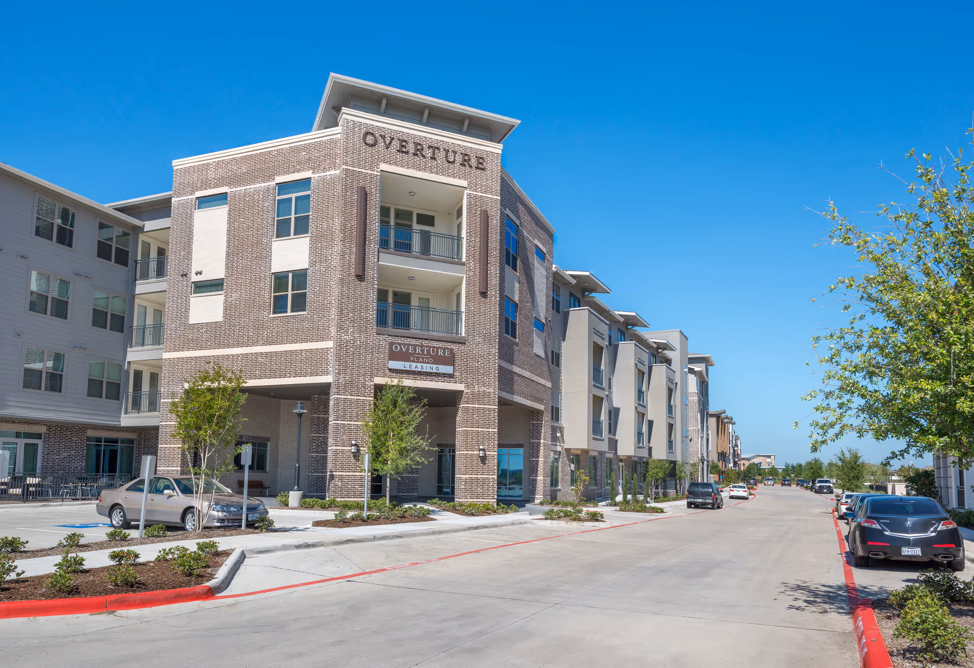 Exterior view of a modern multi-story residential building named Overture Plano with balconies, parked cars along the street, small trees, and clear blue sky.