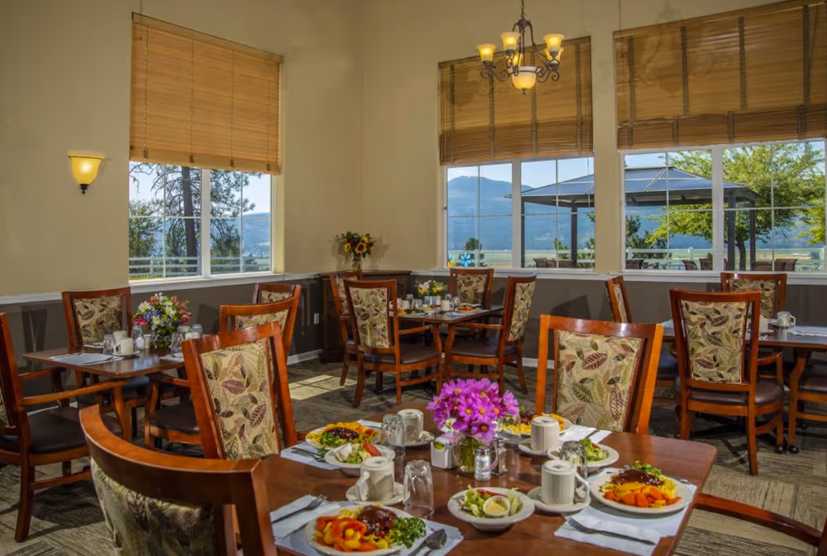 Dining room with wooden tables and chairs featuring floral upholstery. Tables are set with plates of food, cups, and utensils. Large windows with bamboo blinds offer a view of trees, mountains, and an outdoor seating area with a gazebo. A chandelier and wall sconce provide lighting.