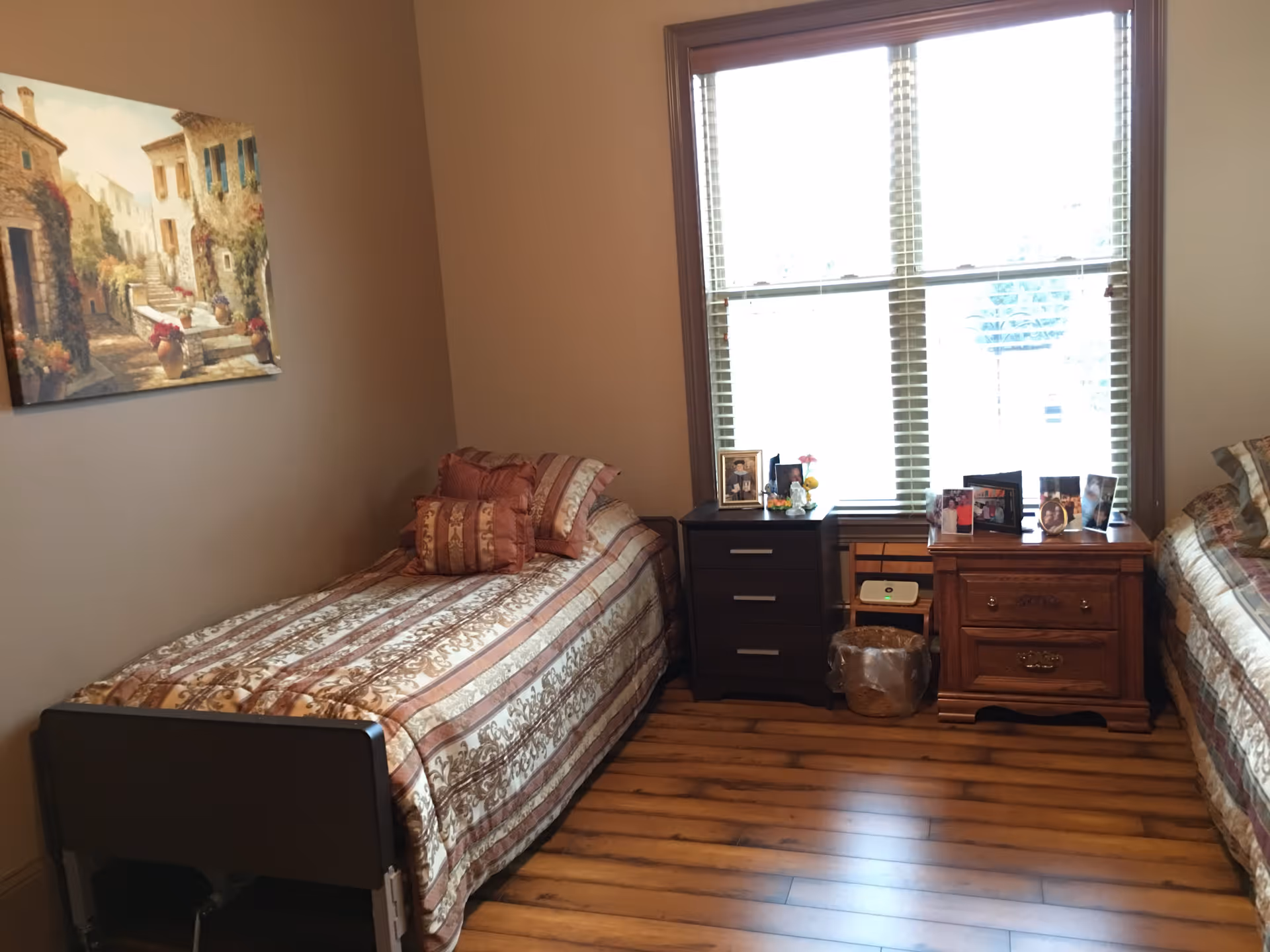 A cozy bedroom with two single beds on either side of a window with blinds. Each bed has a patterned bedspread and multiple pillows. Between the beds are two nightstands, one dark-colored with three drawers and the other wooden with two drawers, both holding framed photos and small decorative items. The floor is wooden, and a painting of a village scene hangs on the left wall.