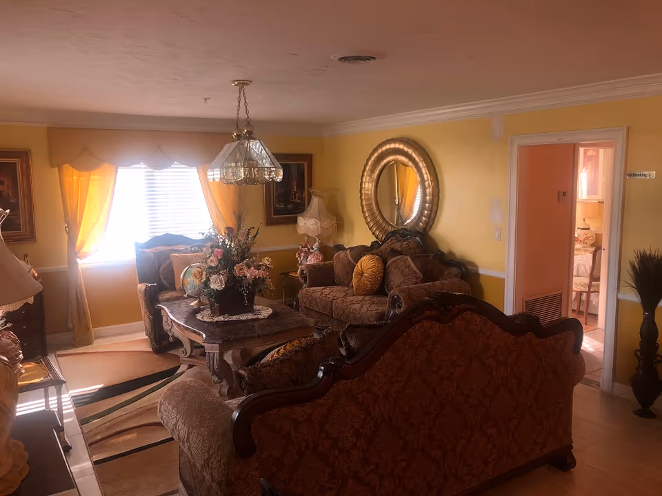 A warmly lit living room with yellow walls and classic furniture including a sofa, loveseat, and armchair with ornate wood detailing. A glass chandelier hangs from the ceiling above a coffee table adorned with a floral arrangement and decorative items. A large round mirror is mounted on the wall, and yellow curtains frame a window letting in natural light. The room has a patterned area rug and a doorway leading to another room with a glimpse of a bed and floral decor.
