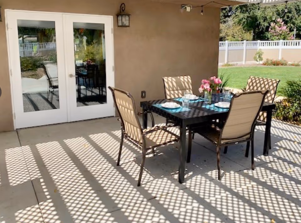 Outdoor patio area with a black metal table set for four, featuring beige cushioned chairs and a vase with pink flowers. The patio has a lattice overhead casting patterned shadows on the concrete floor. White double doors lead inside, and a garden with green grass and plants is visible in the background.