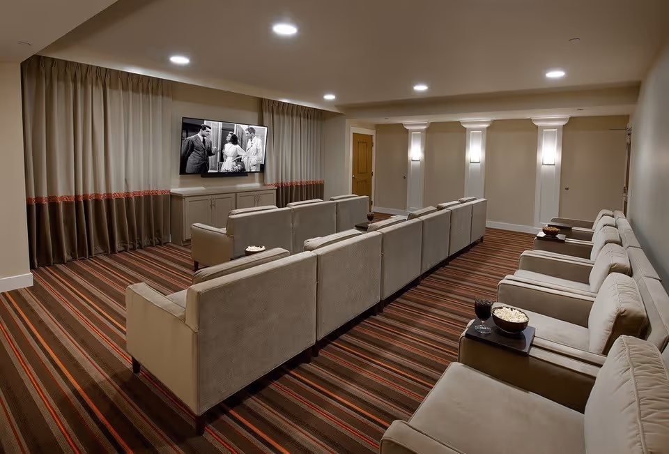 A cozy media room with multiple beige armchairs arranged in rows facing a wall-mounted flat-screen TV playing a black-and-white movie. The room has striped carpet flooring, beige walls with white columns, and soft lighting from wall sconces and recessed ceiling lights. There are small tables between some chairs holding bowls of popcorn and glasses of dark beverages.