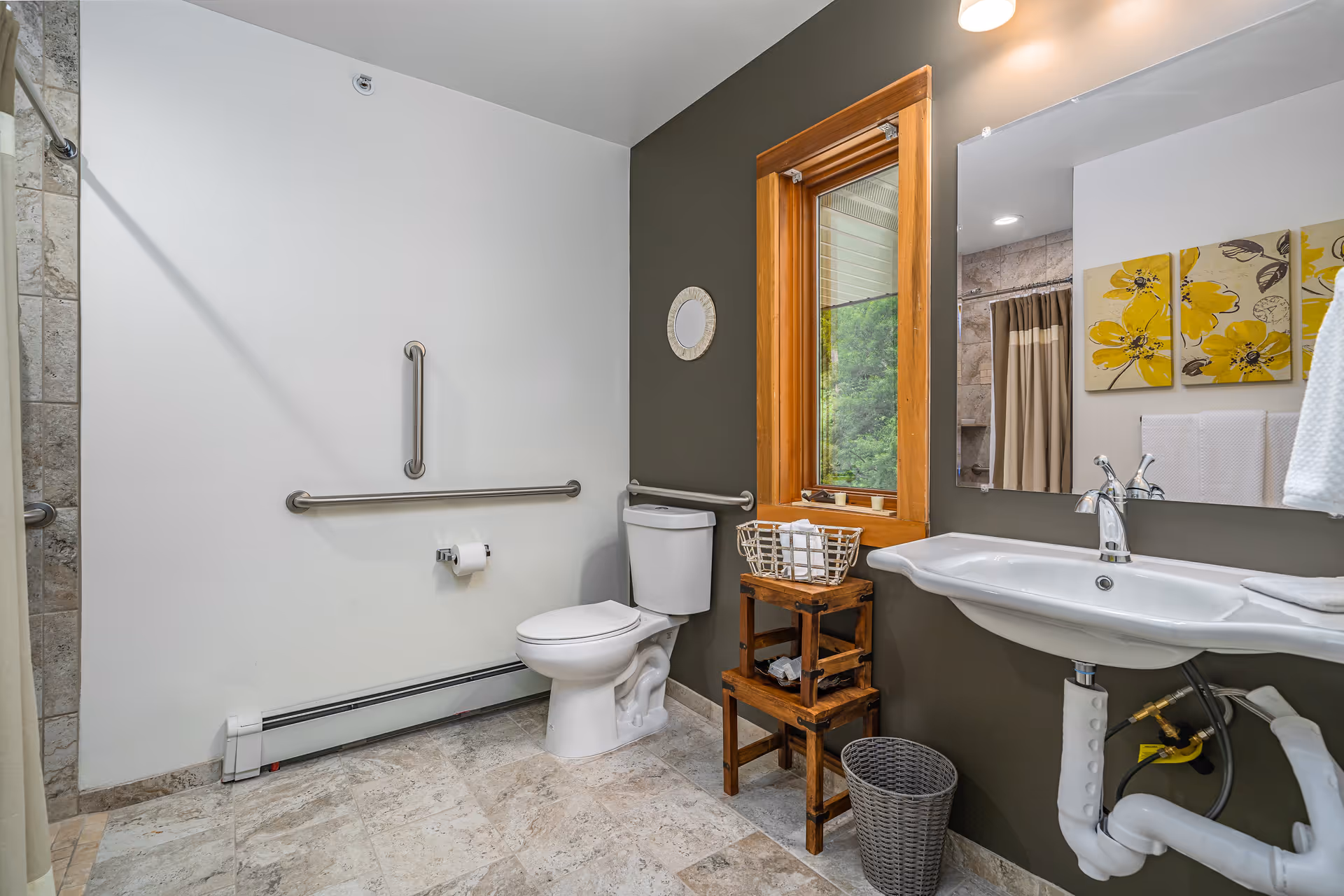 A clean and accessible bathroom featuring a white toilet with grab bars on the adjacent walls, a white sink with exposed plumbing, a wooden-framed window, a small wooden stool with a basket on top, a wastebasket, and a large mirror above the sink. The walls are painted white and dark gray, and there are three yellow floral paintings reflected in the mirror.