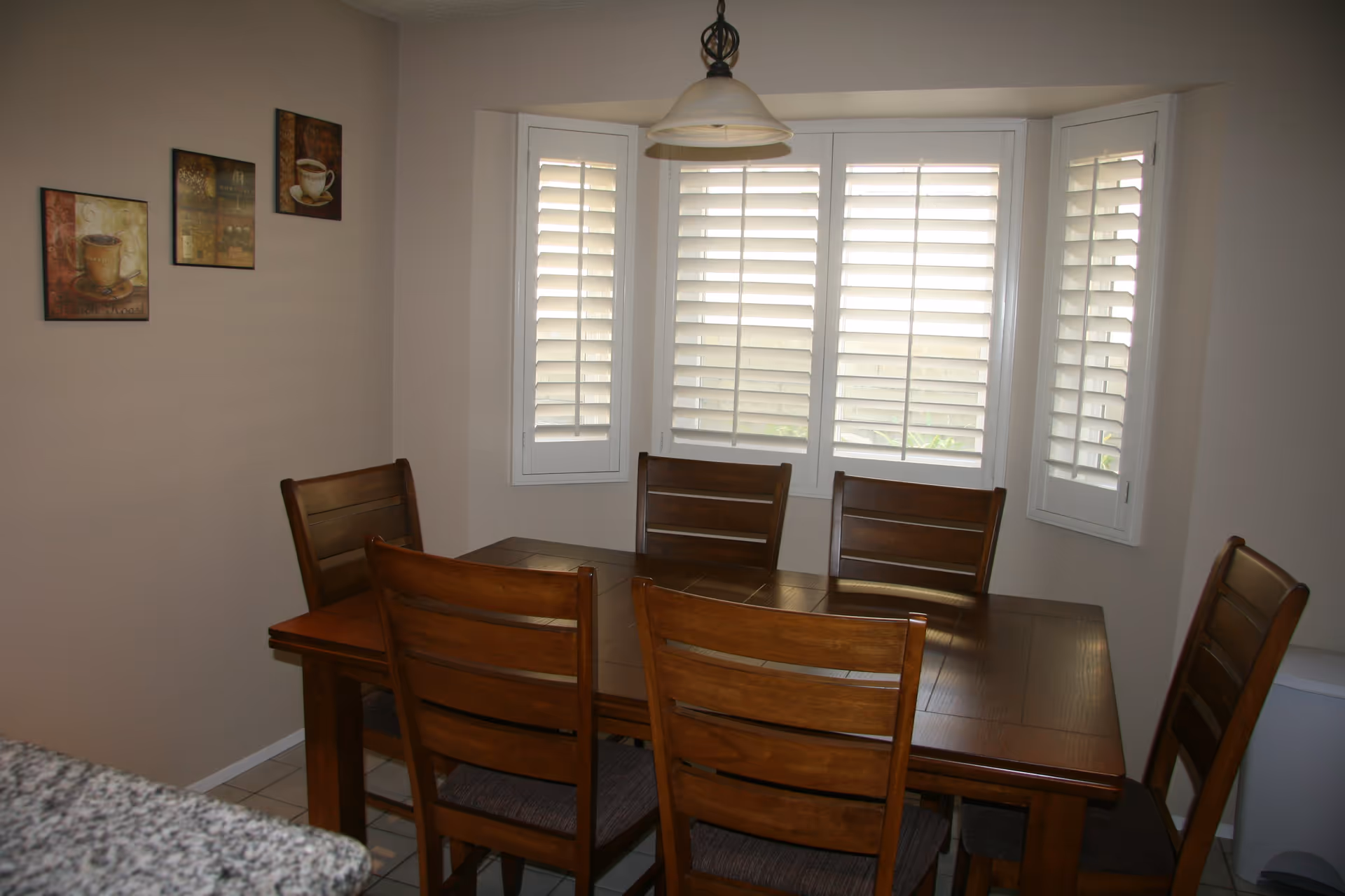 A dining area with a wooden table and six matching wooden chairs with cushioned seats. The room has beige walls with three coffee-themed framed pictures on one wall. There is a large window with white plantation shutters letting in natural light, and a hanging light fixture above the table.