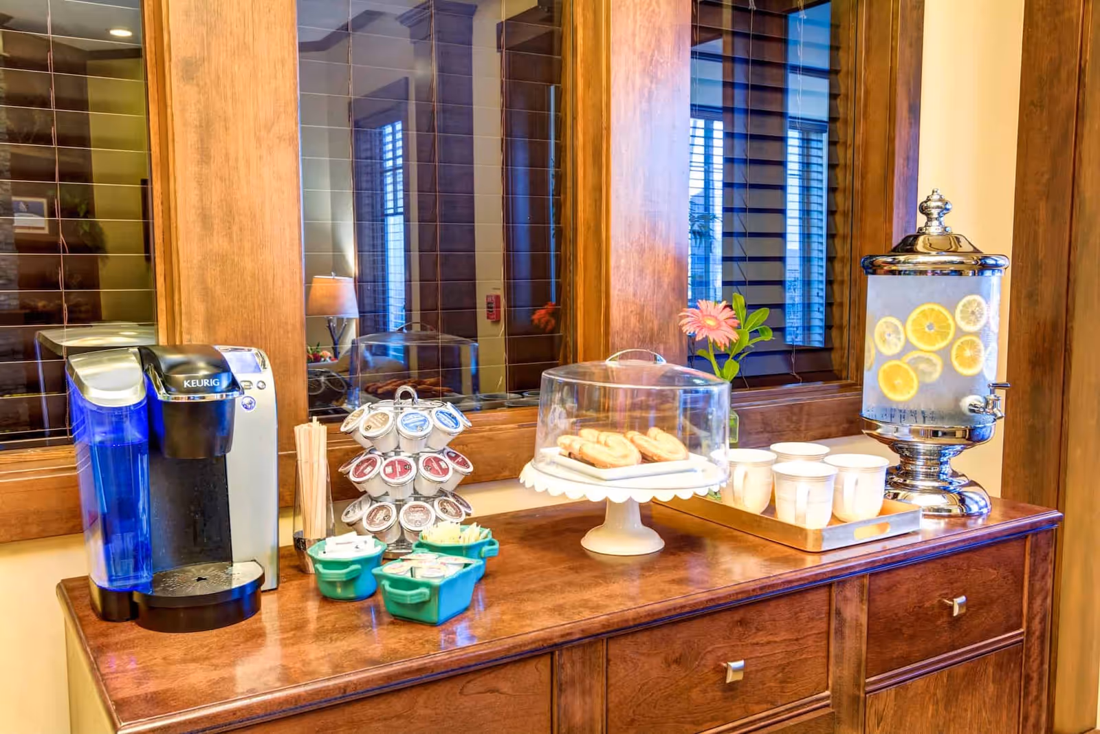A countertop beverage station with a Keurig coffee maker, K-cup rack, a covered plate of pastries, cups on a tray, and a lemon-infused water dispenser in front of wooden windows.