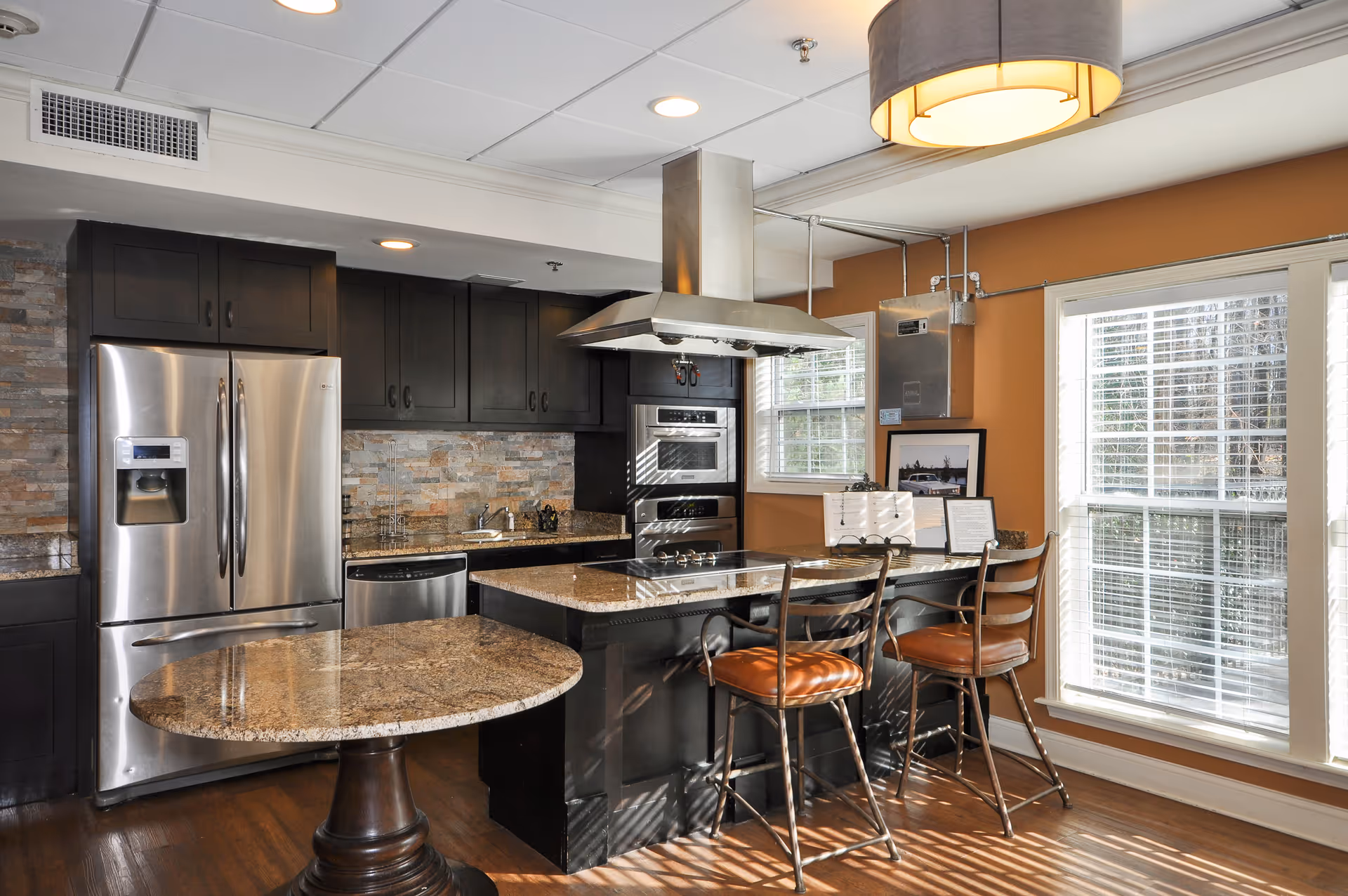 Modern kitchen featuring stainless steel appliances, a granite-topped island with bar stools, and large windows letting in sunlight.