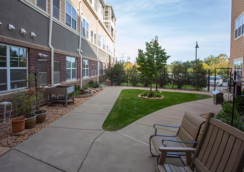 Outdoor courtyard area at a senior living facility with a concrete walkway, green grass, a small tree surrounded by rocks, potted plants along the building, and outdoor seating benches. The building has multiple windows and is three stories tall. A black metal fence encloses the area with trees and a clear sky in the background.