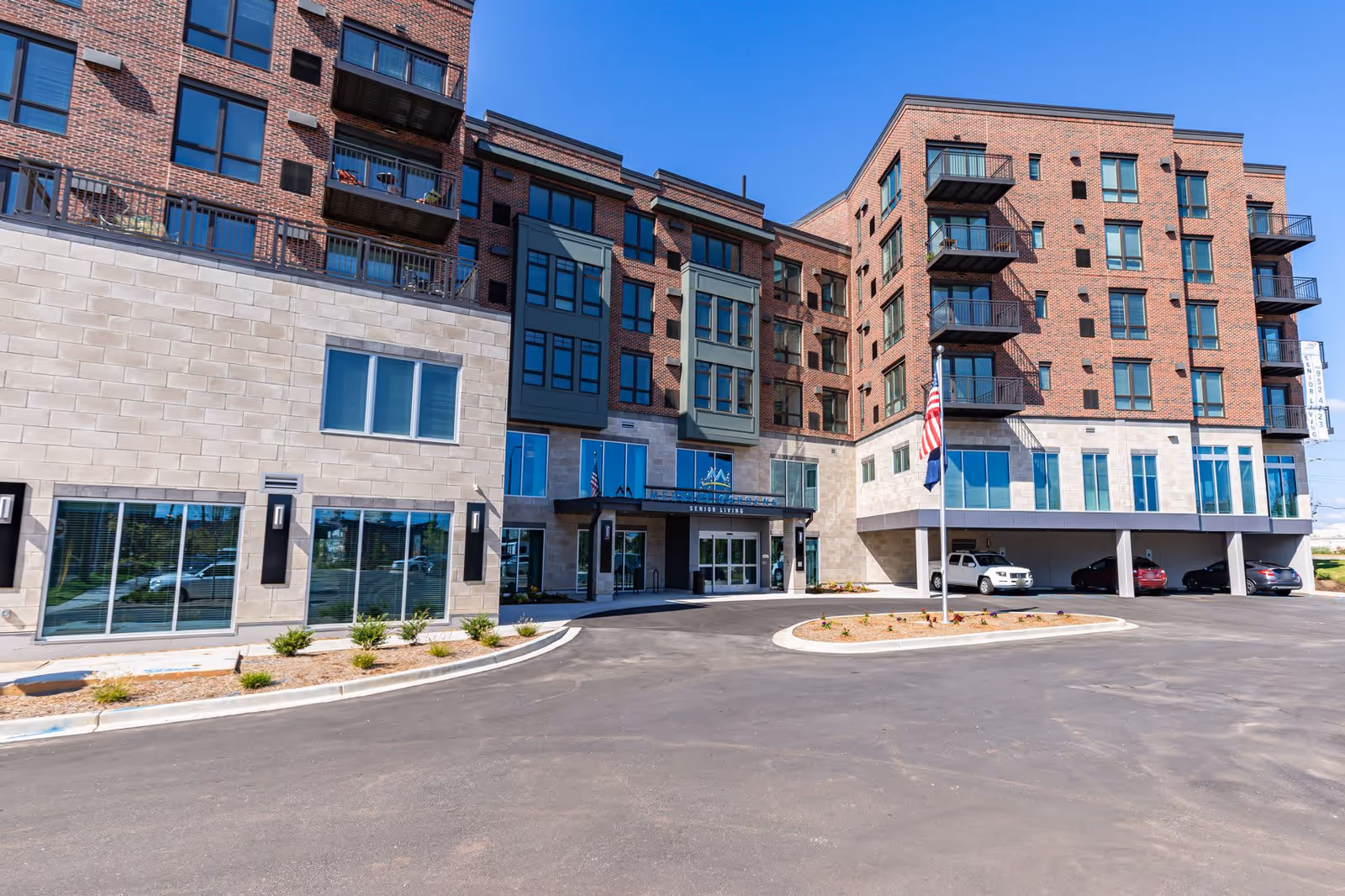 Exterior view of Merrill Gardens at Greenville, a multi-story senior living facility with brick and stone facade, balconies, large windows, an American flag on a flagpole, and a parking area with several cars under a covered section.
