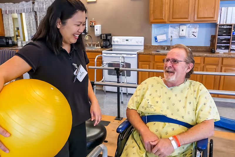A healthcare worker holding a large yellow exercise ball smiles and interacts with a male patient in a wheelchair wearing a hospital gown and a blue belt in a room with kitchen cabinets, a stove, and a sink in the background.