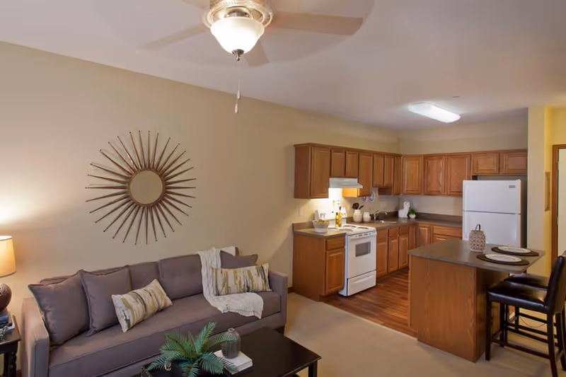 A cozy living area with a gray sofa adorned with patterned pillows and a throw blanket, a black coffee table with decorative plants and books, and a wall-mounted sunburst mirror. Adjacent to the living area is a kitchen featuring wooden cabinets, a white stove, a white refrigerator, and a kitchen island with two black bar stools and place settings. The room is softly lit by a ceiling fan light and a table lamp.