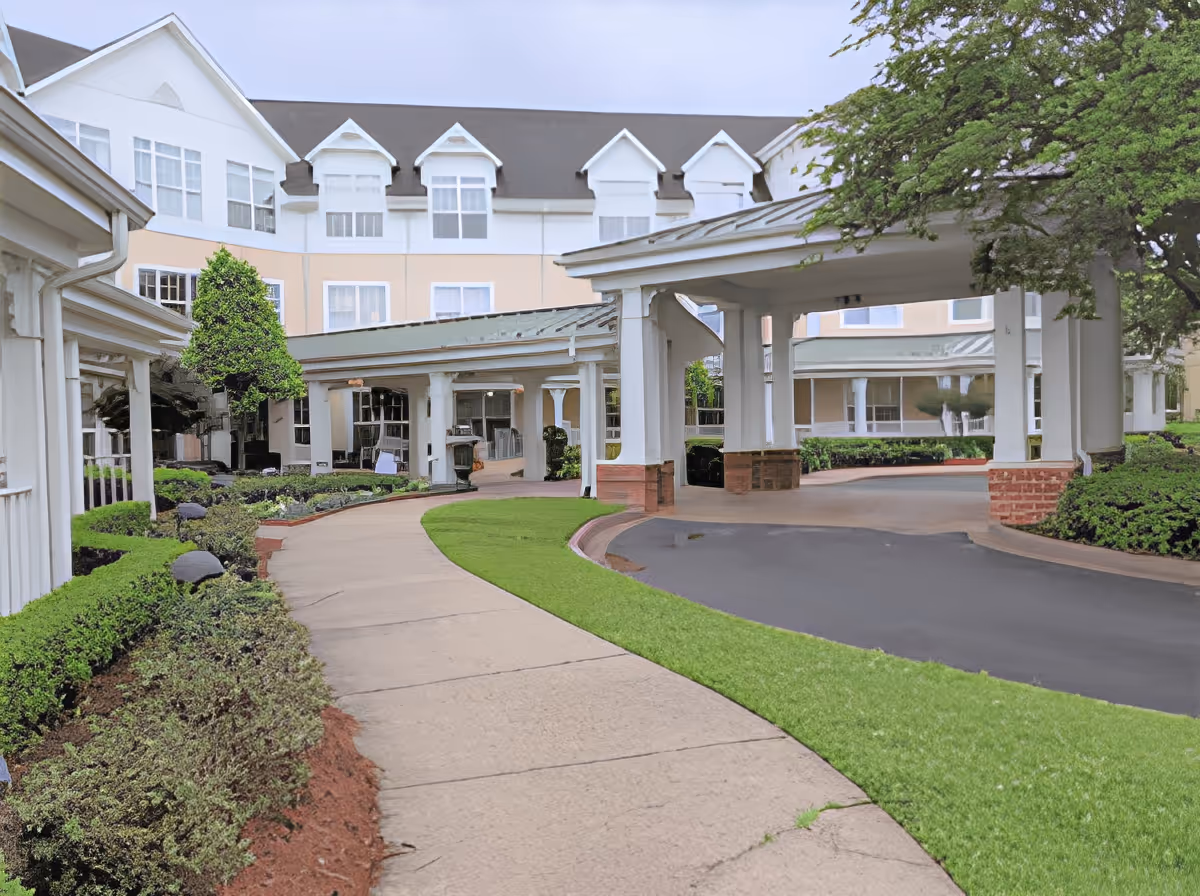 Exterior view of SummerHouse Vista Shores showing a covered driveway entrance with white pillars, a curved sidewalk, well-maintained green lawns, shrubs, and a multi-story building with white and beige walls and multiple windows in the background.