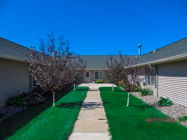 Outdoor courtyard area with a concrete walkway flanked by green grass and small trees, surrounded by single-story beige buildings under a clear blue sky.